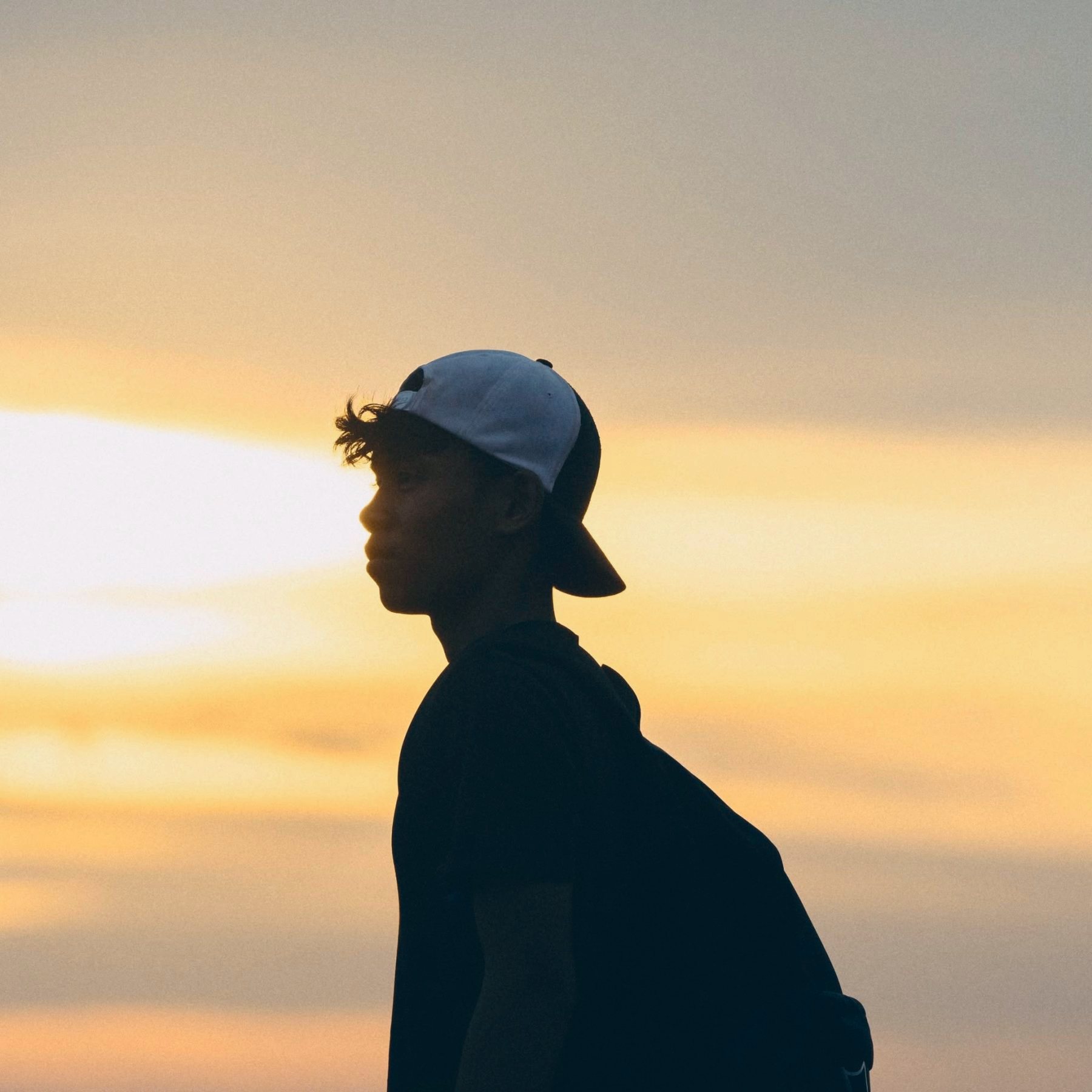 A young man in a cap and backpack, silhouetted against a sunset
