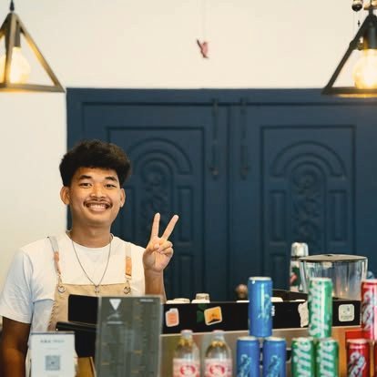 A smiling barista in an apron behind the GP Cafe counter, flashing a peace sign