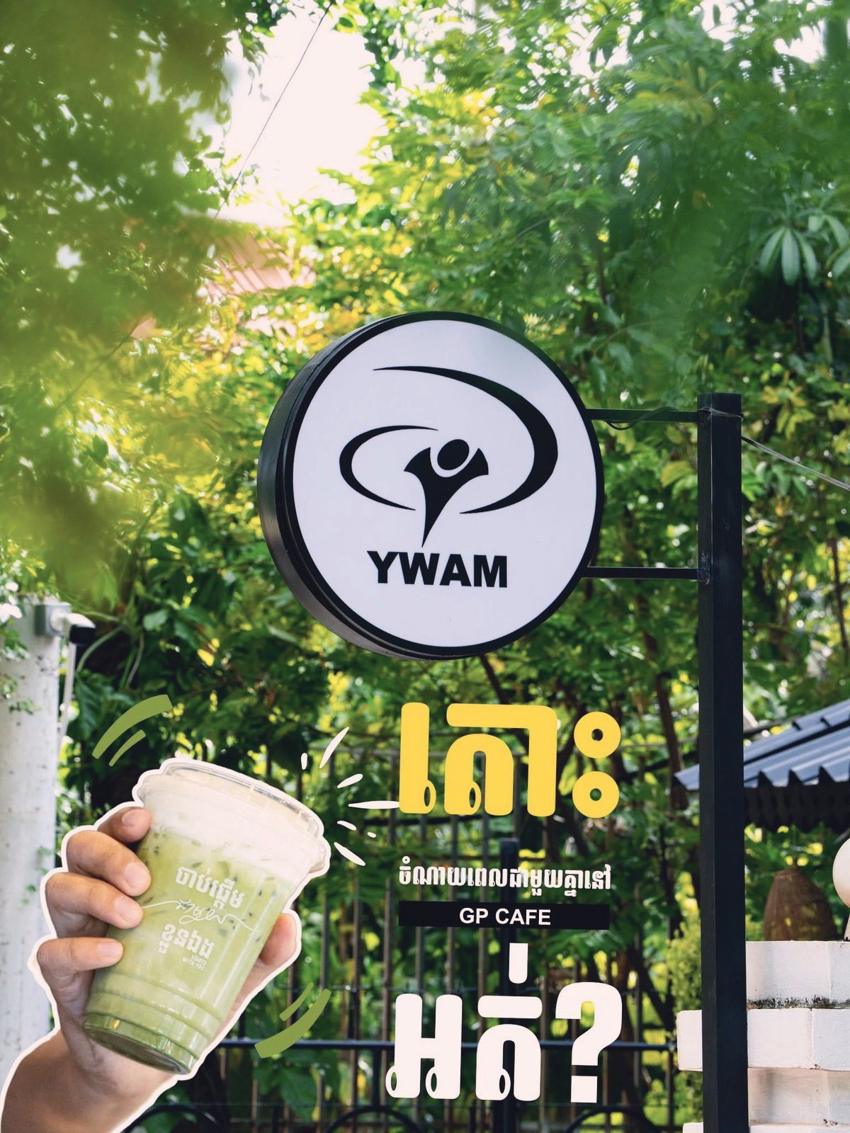 The YWAM sign and the GP Cafe sign hanging among green leaves, with an iced green drink held up in front