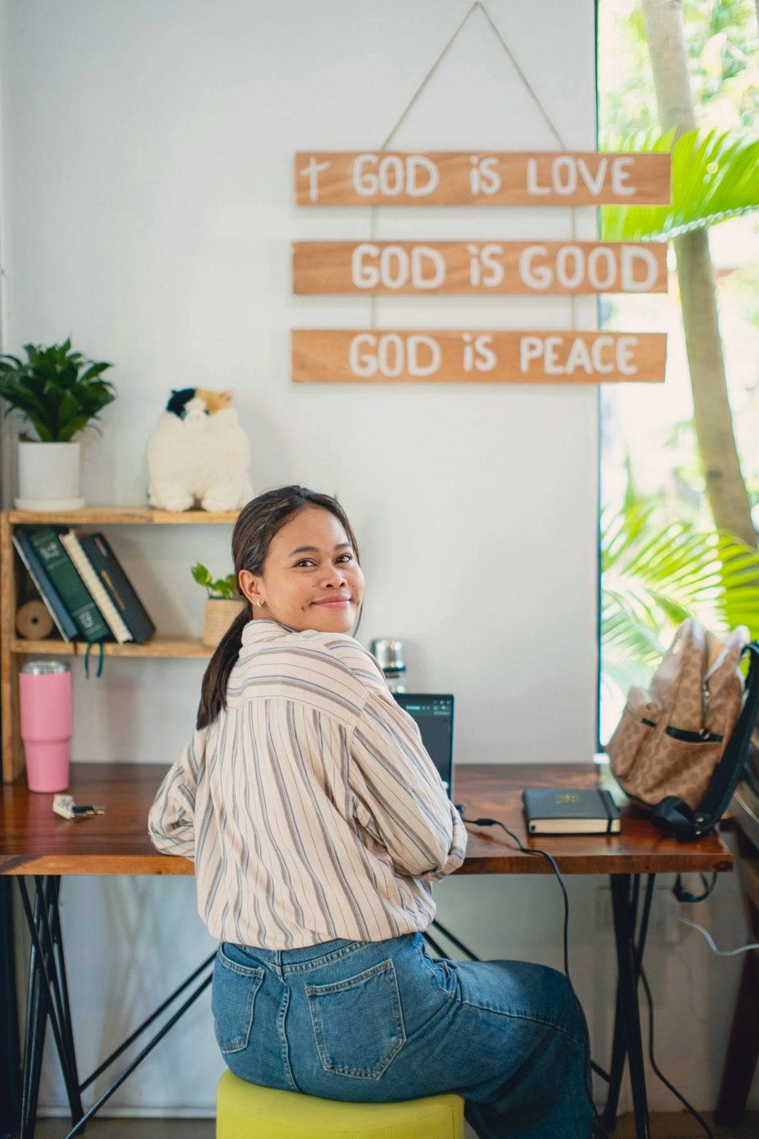 A staff member turning from her desk to smile, under wooden signs reading God is good and God is peace