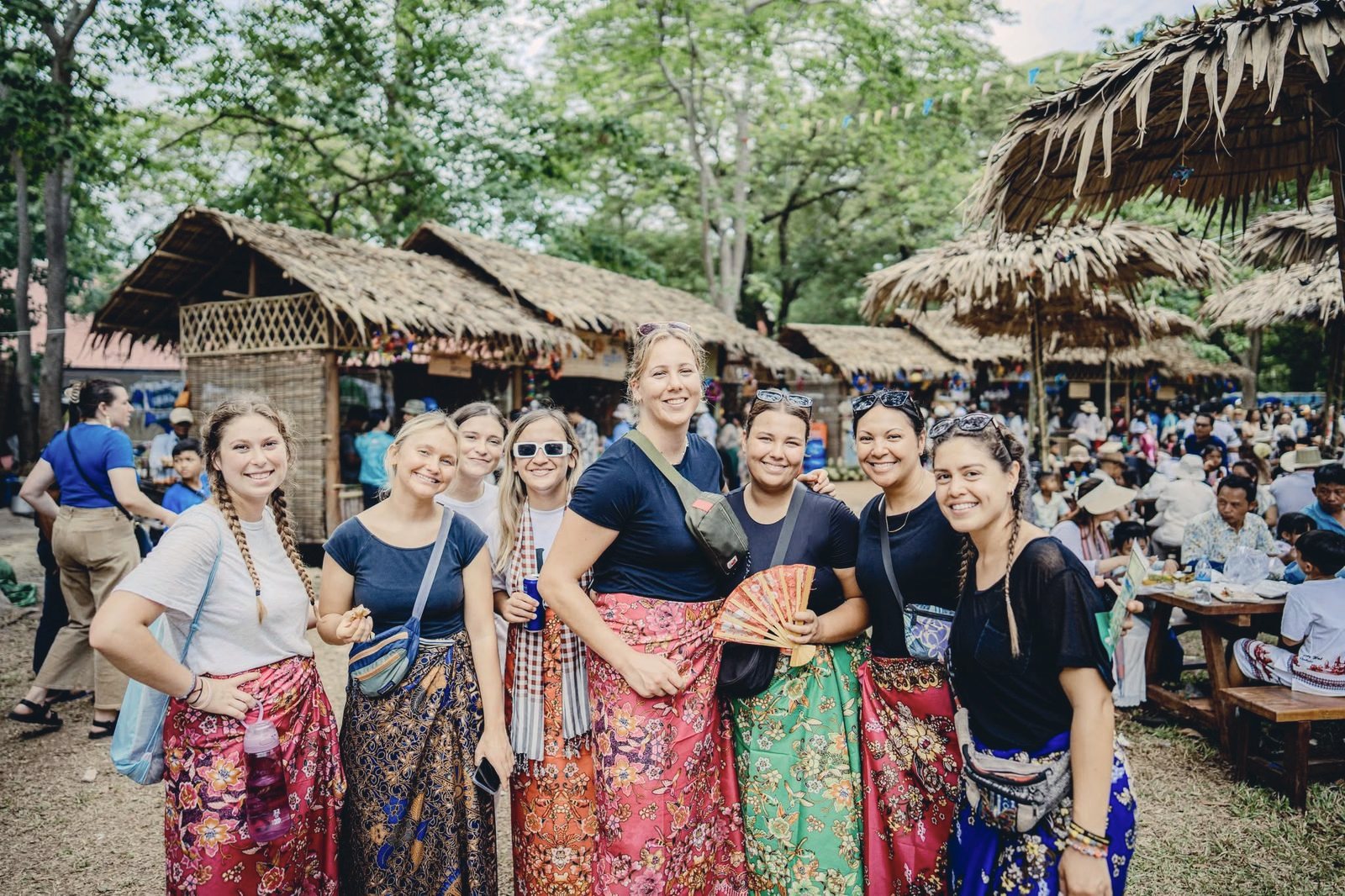 A visiting team in Khmer skirts laughing together at a market of thatched stalls