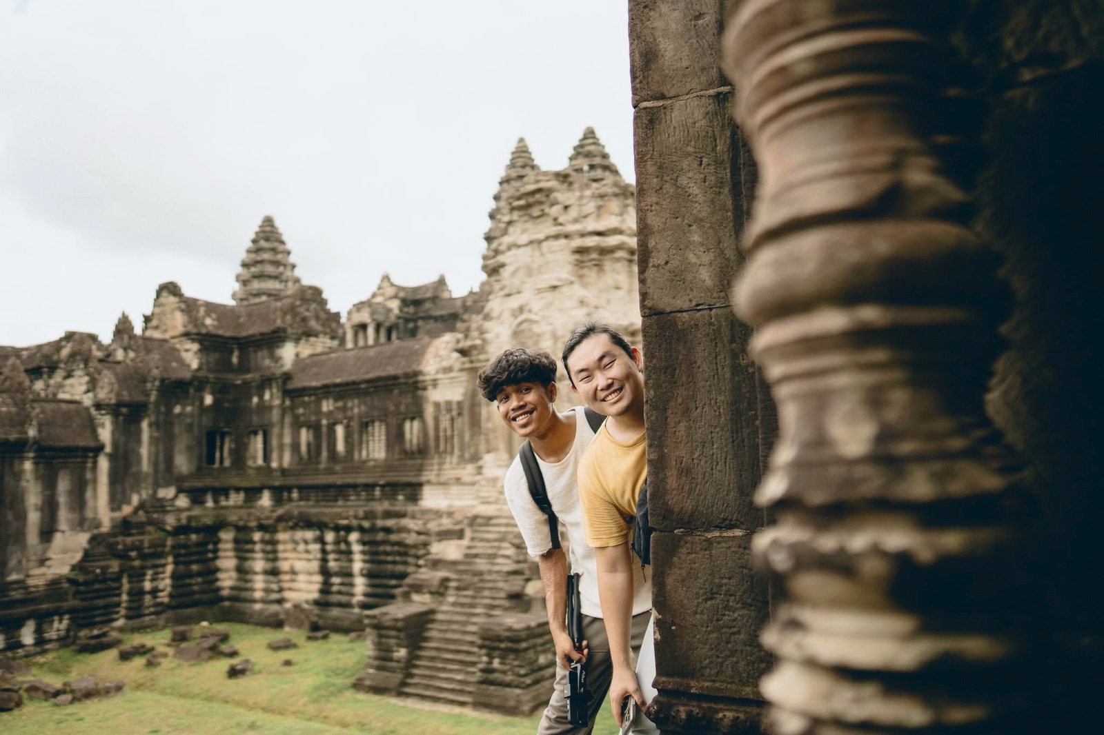 Two young men grinning from behind a stone pillar at Angkor Wat