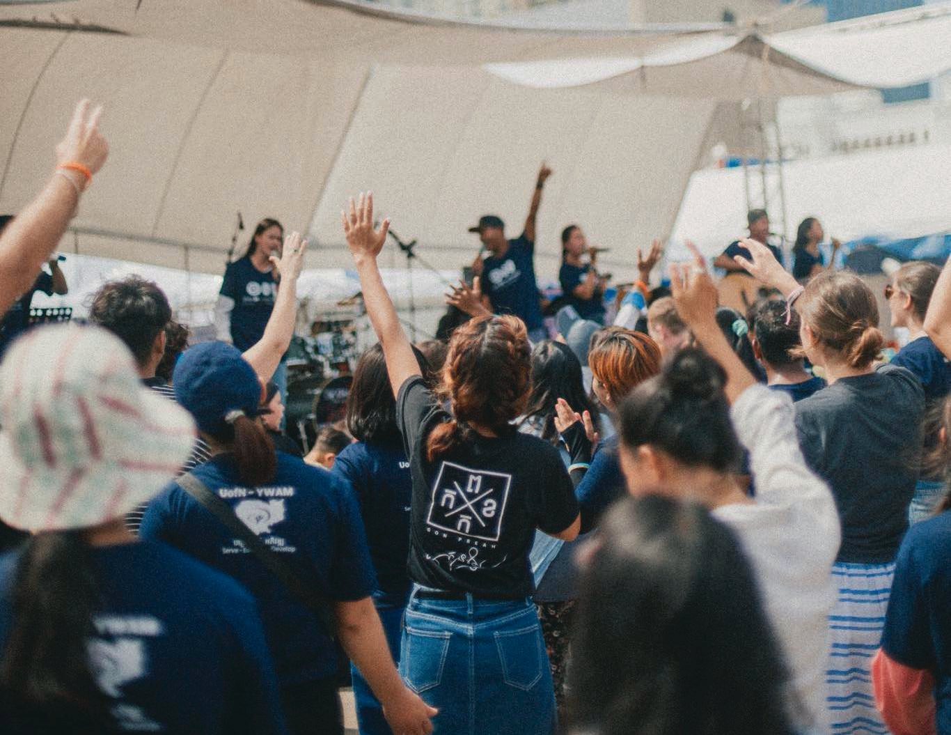 The community worshipping with hands raised in front of the band, one student in a GonPreah shirt