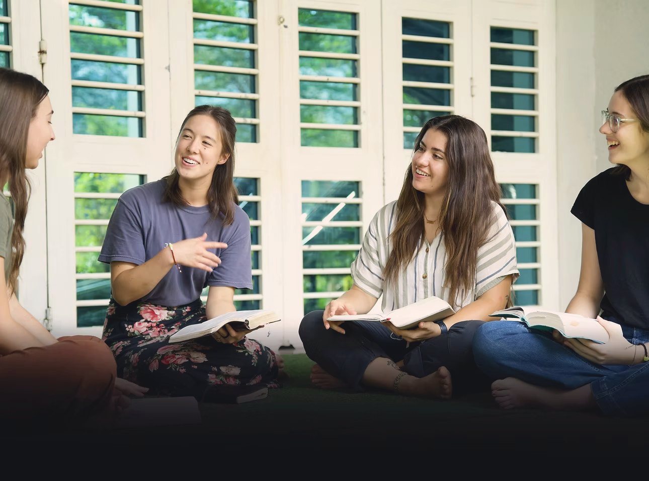 Four students sitting cross-legged on the floor with open Bibles, laughing together in front of louvred windows