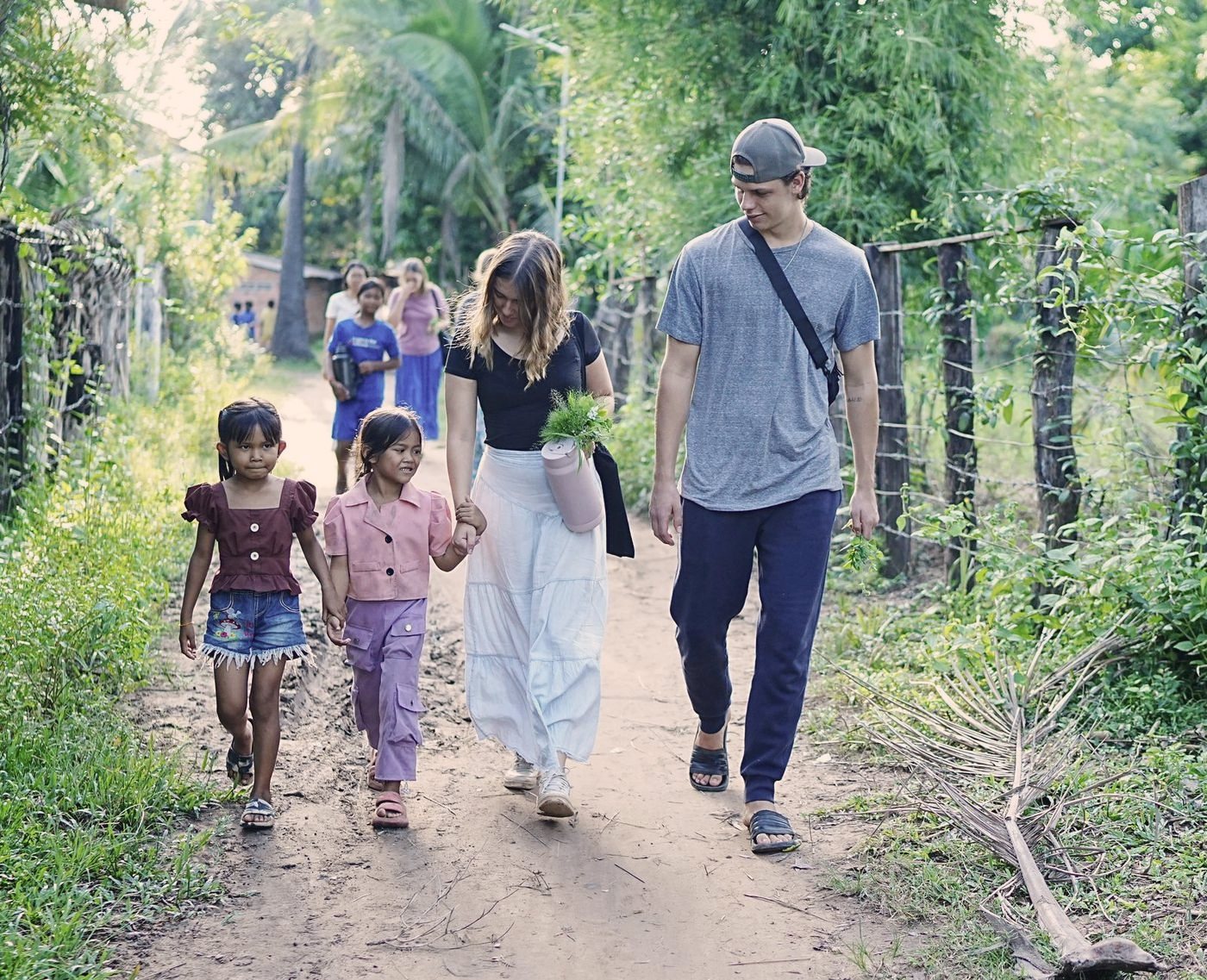 Two DTS students walking down a village path in Cambodia, each holding a young girl's hand