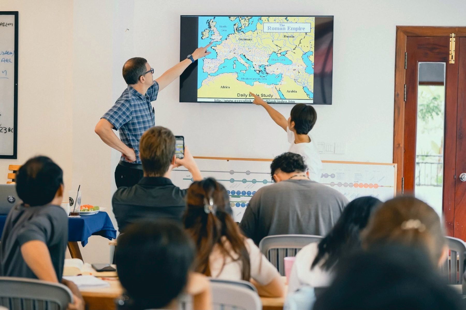A speaker teaching at a map of the world while students listen